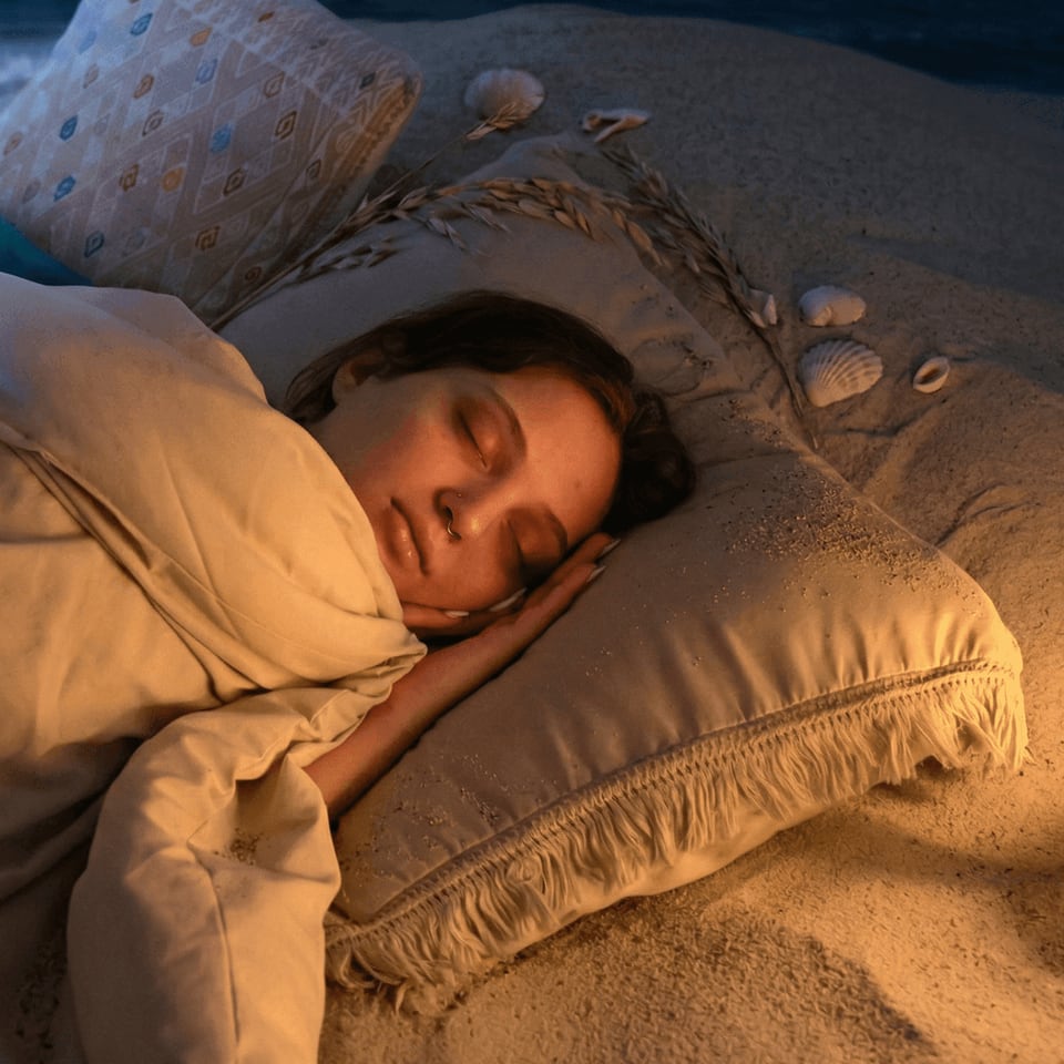 A woman asleep on her side, her ear resting on a fringed pillow in warm light.